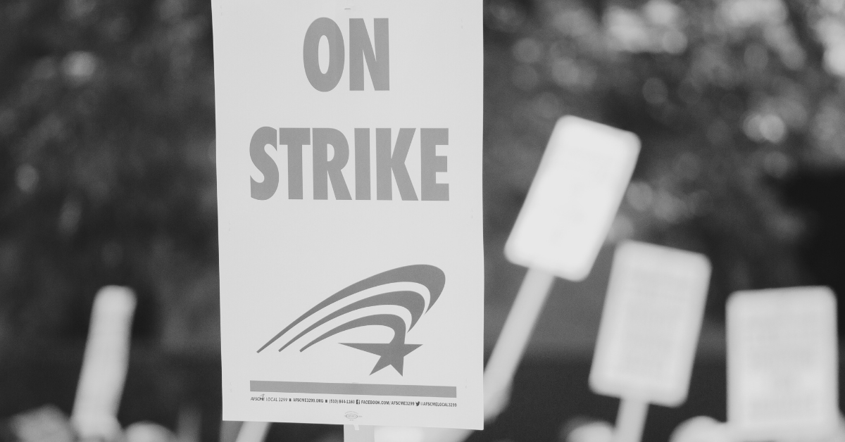 A black and white close-up shows a picket sign that reads "ON STRIKE" in large, bold letters. Below the text is a stylized graphic of a star with five curved lines extending upwards to the left. The sign is held upright, and other blurred picket signs are visible in the background, suggesting a gathering or protest.